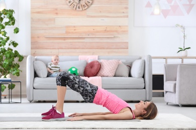 Photo of Young sportive woman doing exercise while her son sitting on sofa at home. Fitness training