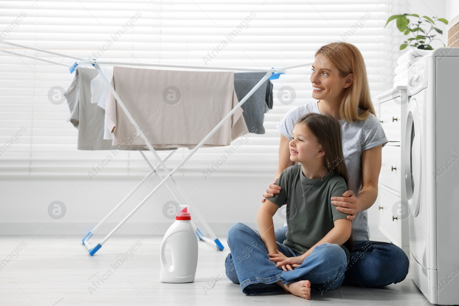 Photo of Mother and daughter sitting on floor near fabric softener and clothes dryer in bathroom, space for text