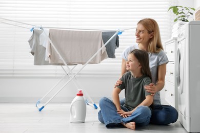 Photo of Mother and daughter sitting on floor near fabric softener and clothes dryer in bathroom, space for text