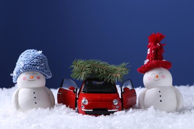 Photo of Cute decorative snowmen and toy car with fir tree branches on artificial snow against blue background