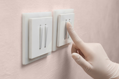 Woman in protective gloves pressing button of light switch indoors, closeup