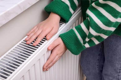 Photo of Girl warming hands on heating radiator indoors, closeup