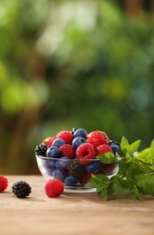 Glass bowl with different fresh ripe berries and mint on wooden table outdoors