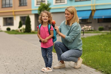 Photo of Happy mother putting apple into with her daughter's backpack near kindergarten outdoors