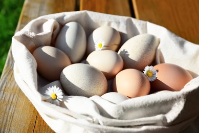 Photo of Sack of assorted eggs and chamomile flowers on wooden table outdoors, closeup