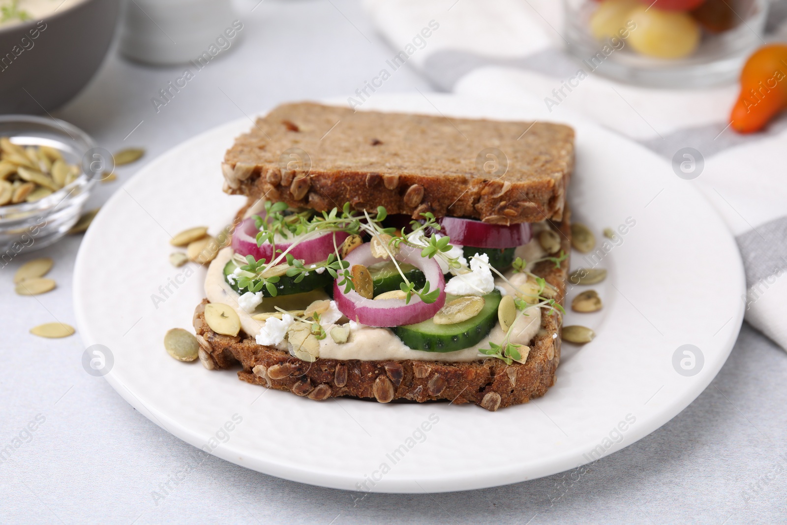 Photo of Tasty vegan sandwich with cucumber, onion, hummus and pumpkin seeds on light grey table