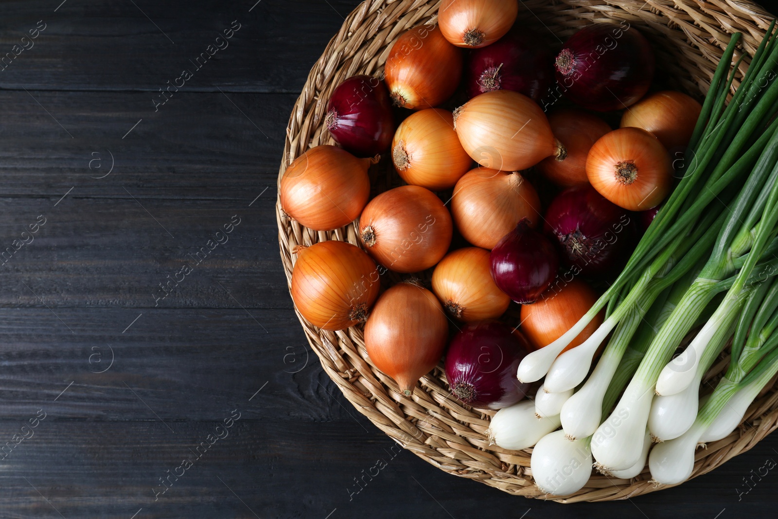 Photo of Wicker mat with different kinds of onions on black wooden table, top view. Space for text