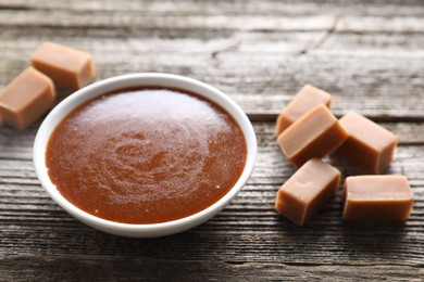 Salted caramel sauce in bowl and yummy candies on wooden table, closeup