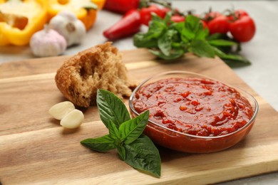 Delicious adjika sauce in glass bowl with bread and ingredients on wooden board, closeup
