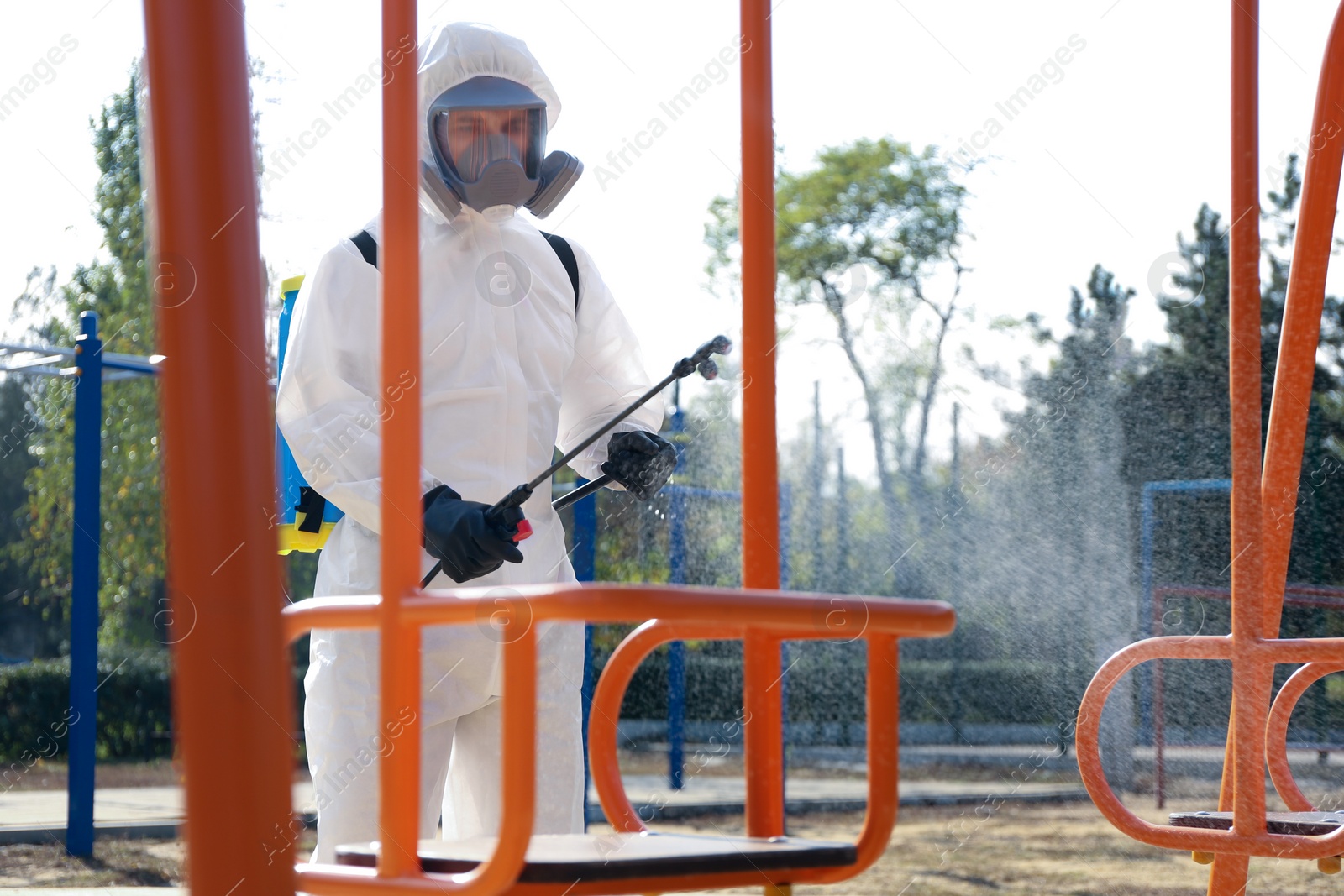 Photo of Man in hazmat suit spraying disinfectant onto swing at children's playground. Surface treatment during coronavirus pandemic