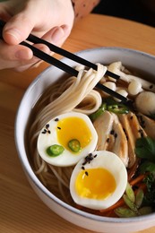 Woman eating delicious ramen with chopsticks at wooden table, closeup. Noodle soup