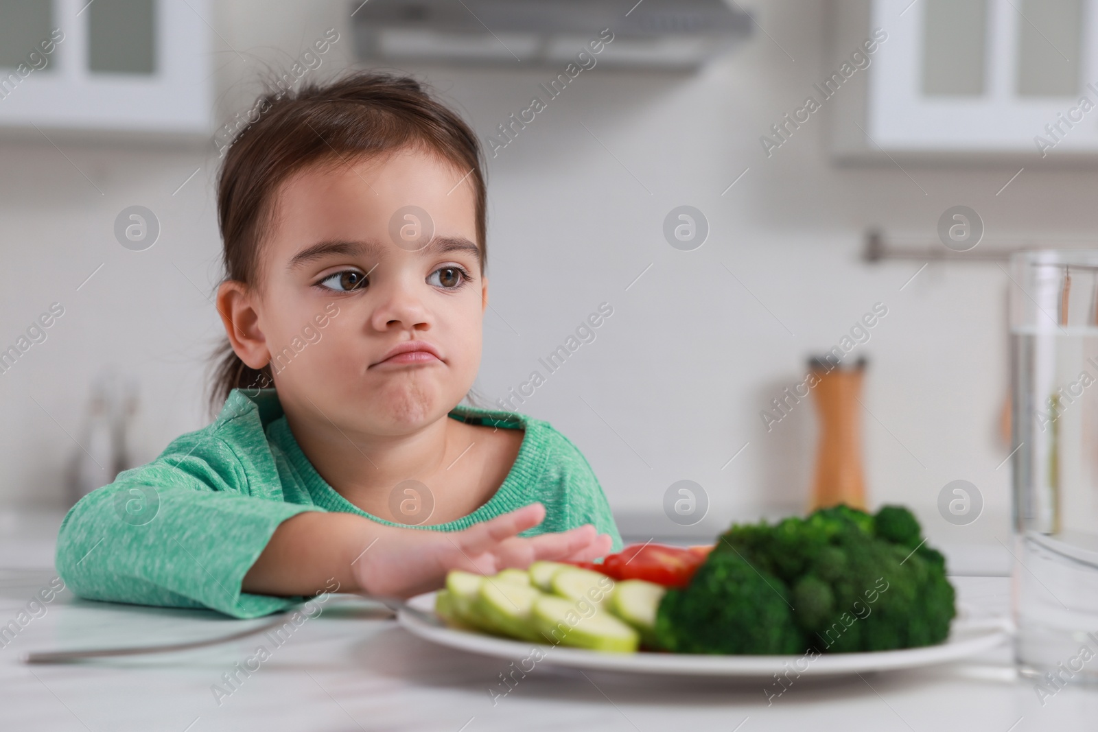 Photo of Cute little girl refusing to eat vegetables in kitchen