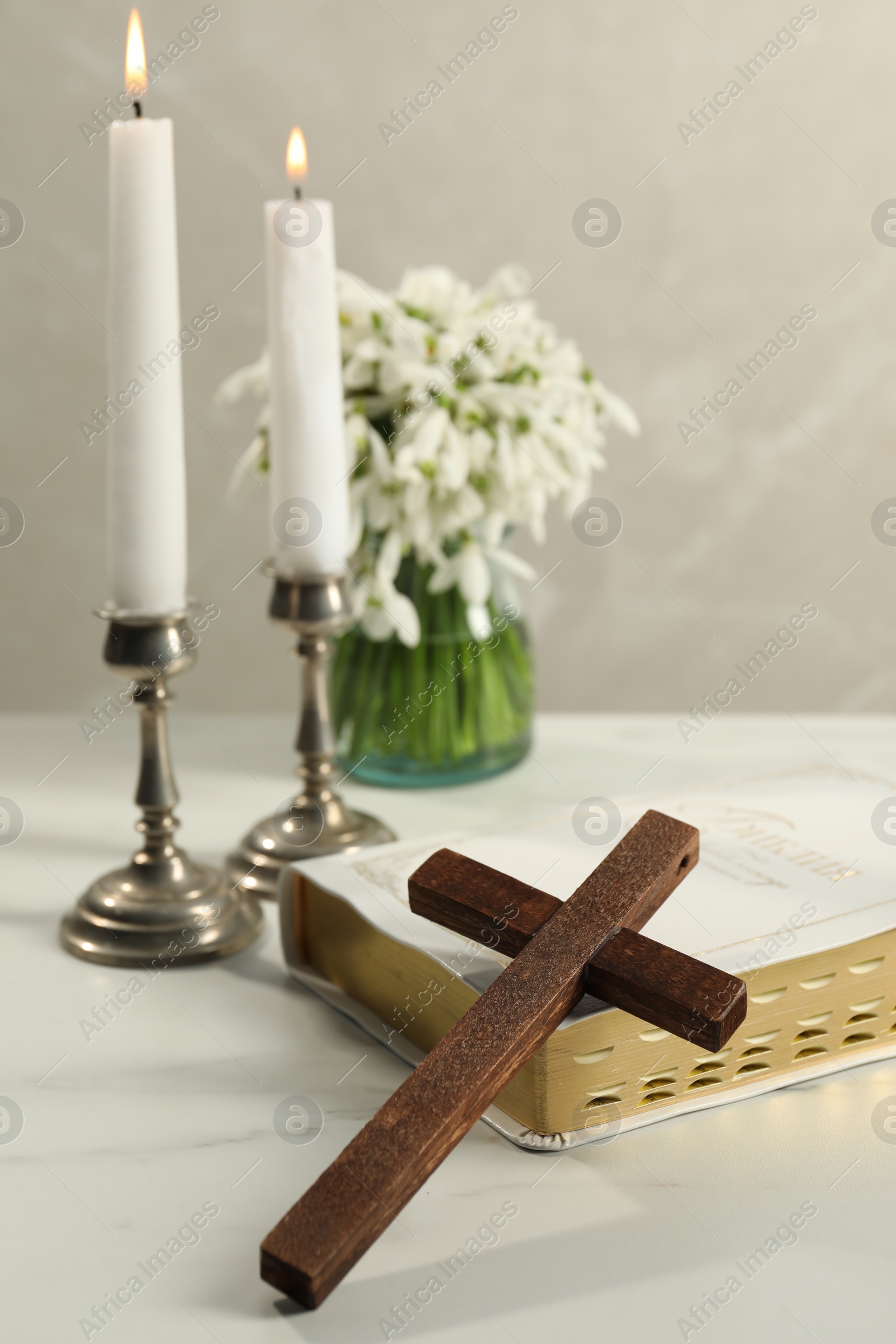 Photo of Burning church candles, wooden cross, Bible and flowers on white marble table