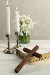 Burning church candles, wooden cross, Bible and flowers on white marble table