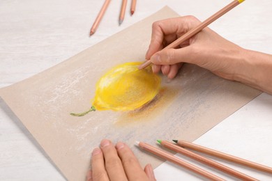 Photo of Woman drawing lemon on paper with pastel pencil at white wooden table, closeup