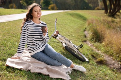 Young woman sitting on green grass and holding cup of coffee near bicycle in park, space for text