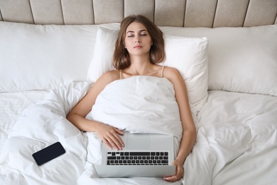 Tired woman with laptop and smartphone sleeping in bed at home, above view