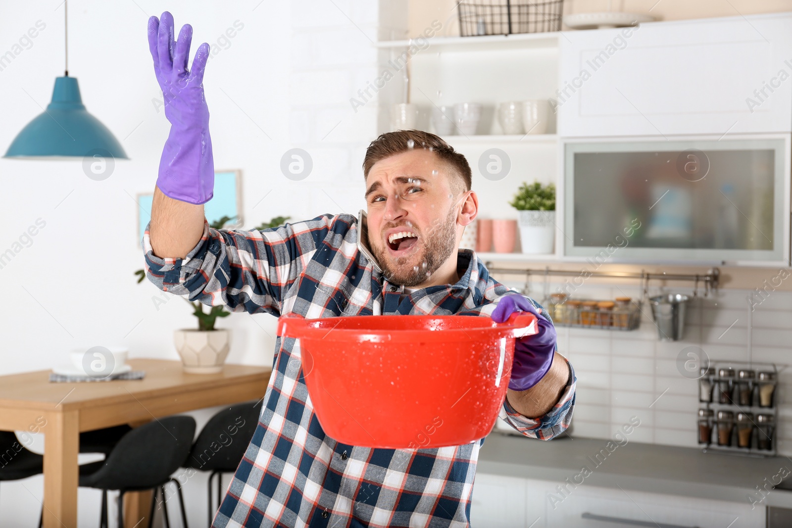 Photo of Emotional young man calling plumber while collecting water leakage from ceiling in kitchen