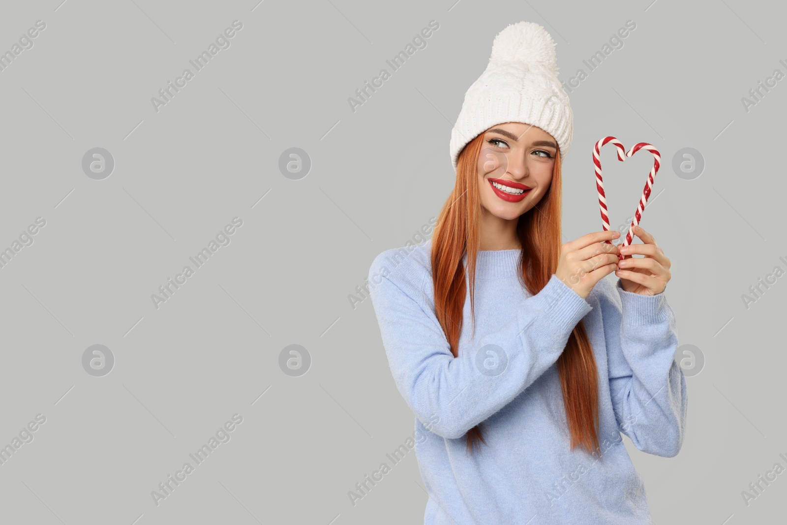 Photo of Young woman in hat and sweater with candy canes on light grey background, space for text. Christmas celebration