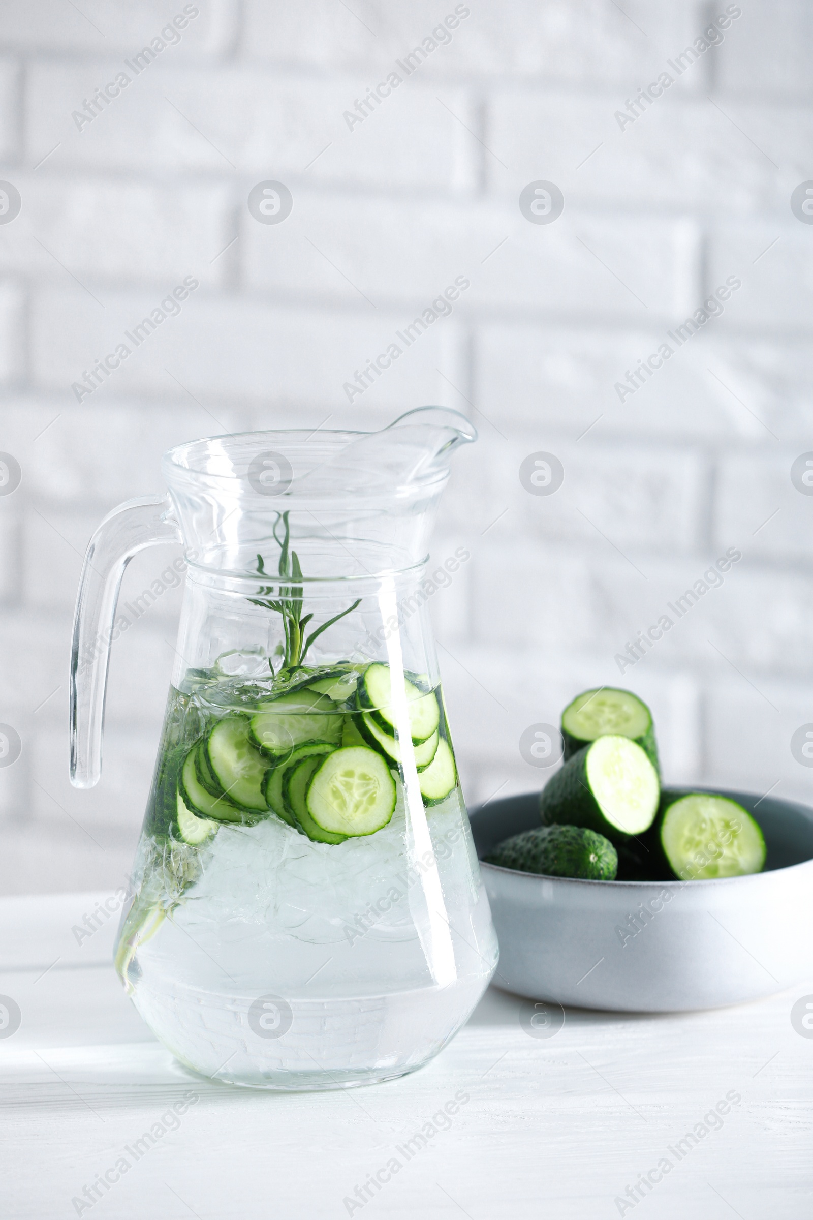 Photo of Refreshing cucumber water with rosemary in jug and vegetables on white table against brick wall