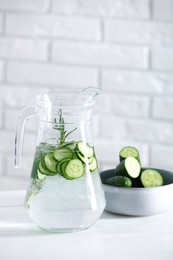 Refreshing cucumber water with rosemary in jug and vegetables on white table against brick wall