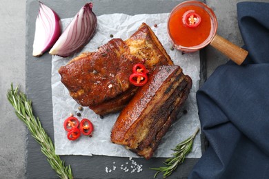 Photo of Pieces of baked pork belly served with sauce, rosemary and chili pepper on grey table, top view