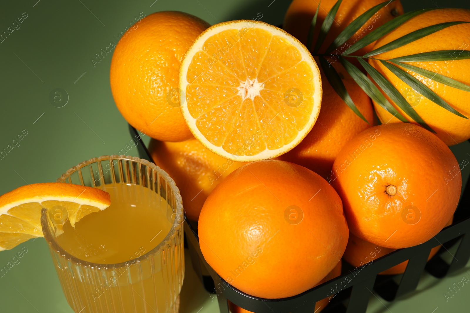 Photo of Fresh oranges in metal basket and glass of juice on green background, closeup