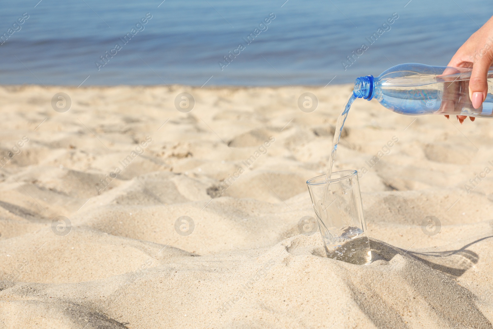 Photo of Woman pouring refreshing drink into glass on sandy beach, space for text. Hot summer day
