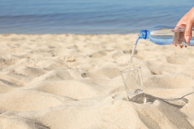 Photo of Woman pouring refreshing drink into glass on sandy beach, space for text. Hot summer day