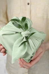 Photo of Furoshiki technique. Woman holding gift packed in green fabric and decorated with ruscus branch, closeup