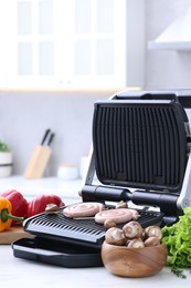 Photo of Electric grill with homemade sausages, mushrooms, and vegetables on white table in kitchen