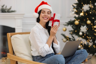 Photo of Celebrating Christmas online with exchanged by mail presents. Smiling woman in Santa hat with gift box during video call on laptop at home