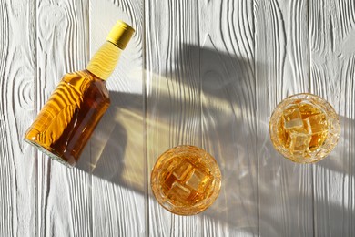Photo of Whiskey with ice cubes in glasses and bottle on white wooden table, flat lay
