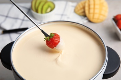 Dipping strawberry into pot with white chocolate fondue on table, closeup