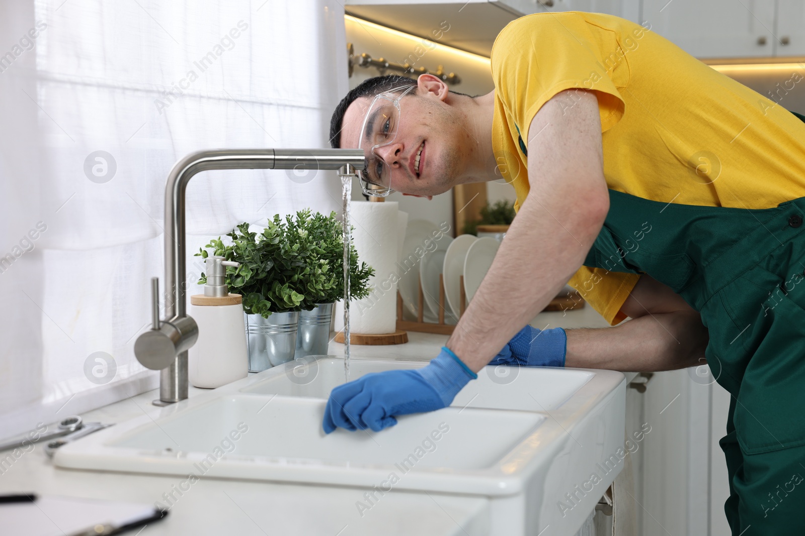 Photo of Smiling plumber wearing protective glasses and gloves examining faucet in kitchen