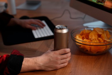 Photo of Young man with energy drink and chips playing video game at wooden desk indoors, closeup