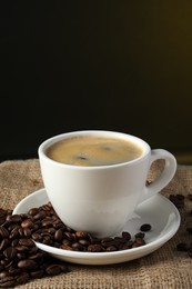 Photo of Cup of hot aromatic coffee and roasted beans on sackcloth against dark background