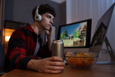 Young man with energy drink and headphones playing video game at wooden desk indoors, focus on can