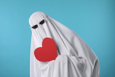 Photo of Cute ghost. Person covered with white sheet in sunglasses holding red heart on light blue background