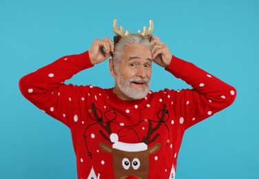 Photo of Senior man in Christmas sweater and reindeer headband on light blue background