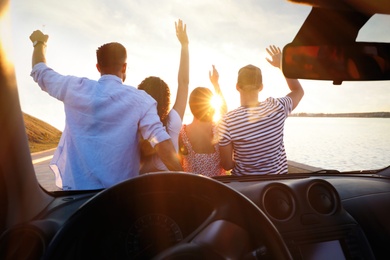 Group of friends near car outdoors at sunset, view through windshield. Summer trip