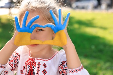 Photo of Little girl making heart with her hands painted in Ukrainian flag colors outdoors, space for text. Love Ukraine concept