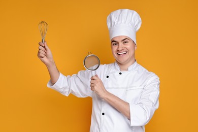 Photo of Portrait of happy confectioner in uniform holding whisk and sieve on orange background