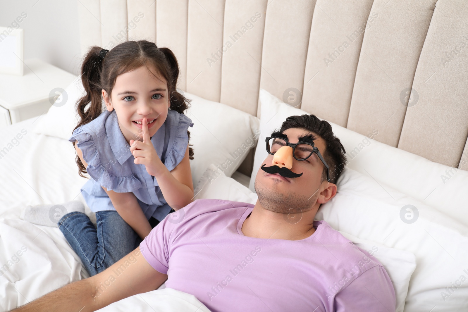 Photo of Cute little girl putting funny glasses on father while he sleeping in bed at home