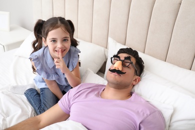 Photo of Cute little girl putting funny glasses on father while he sleeping in bed at home
