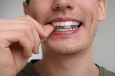 Young man applying whitening strip on his teeth against light grey background, closeup