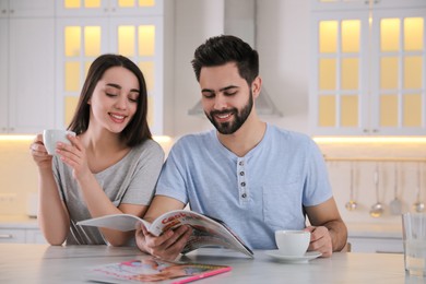 Young couple with cups of drink reading magazine at table in kitchen