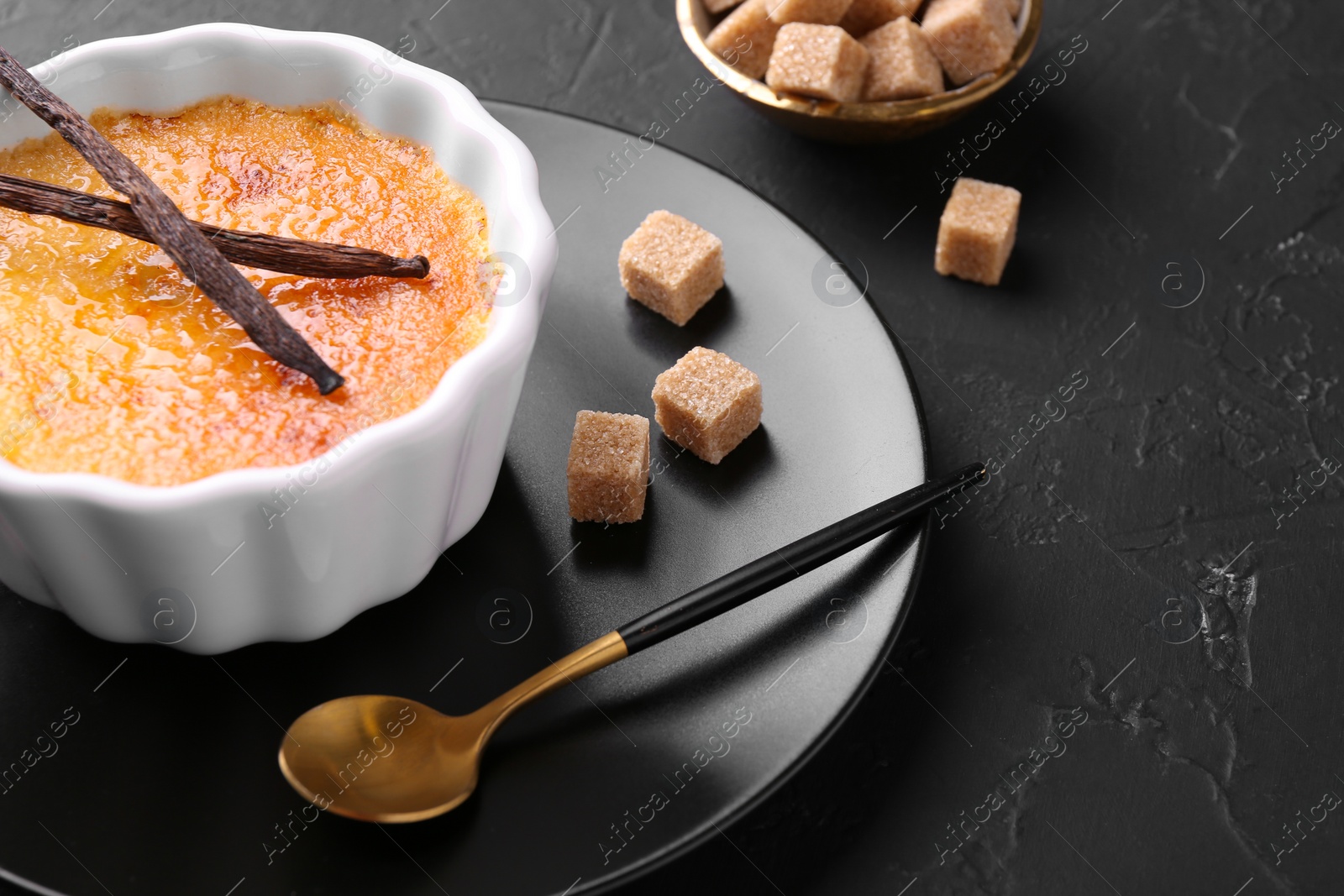 Photo of Delicious creme brulee in bowl, vanilla pods, sugar cubes and spoon on dark gray textured table, closeup