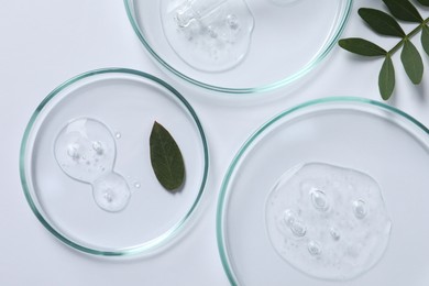 Photo of Petri dishes with samples of cosmetic oil and green leaves on white background, flat lay