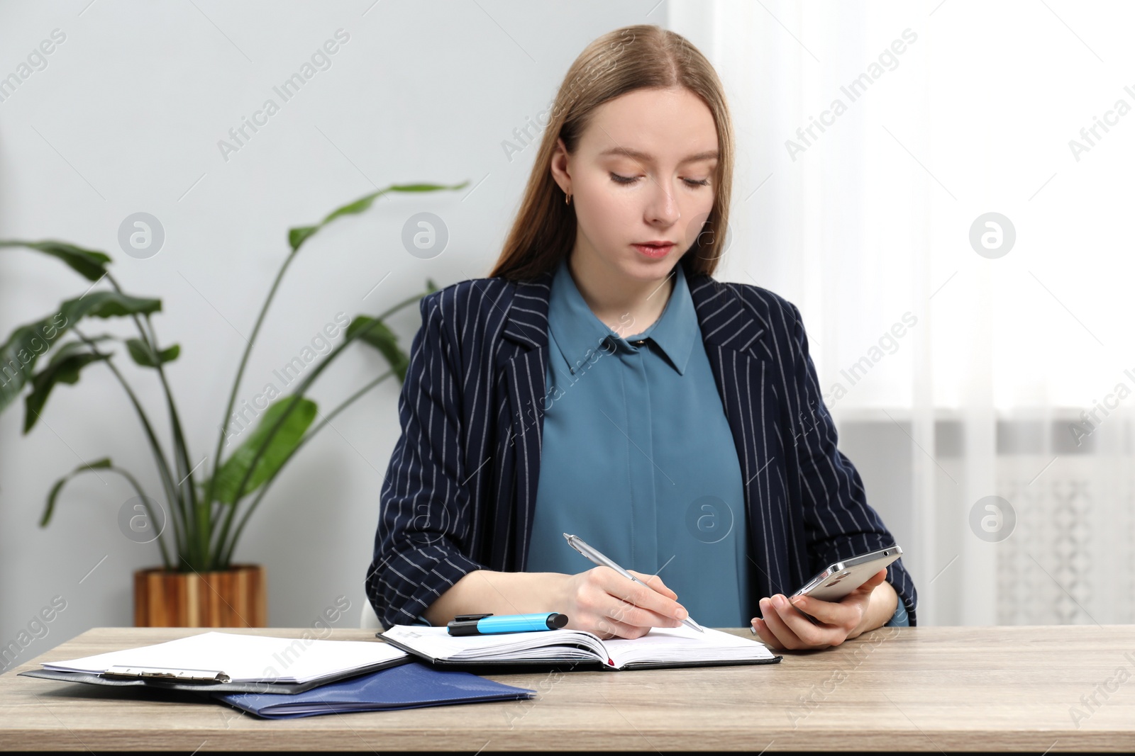 Photo of Woman taking notes while using smartphone at wooden table in office